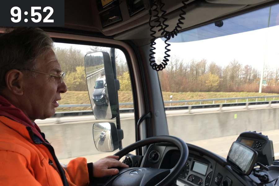 Side view of a truck driver in the cab while driving, with the time stamp 9:52 a.m.