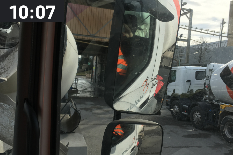 Side view from a concrete mixer truck of the rear view mirrors and a concrete plant with the time stamp 10:07 a.m.