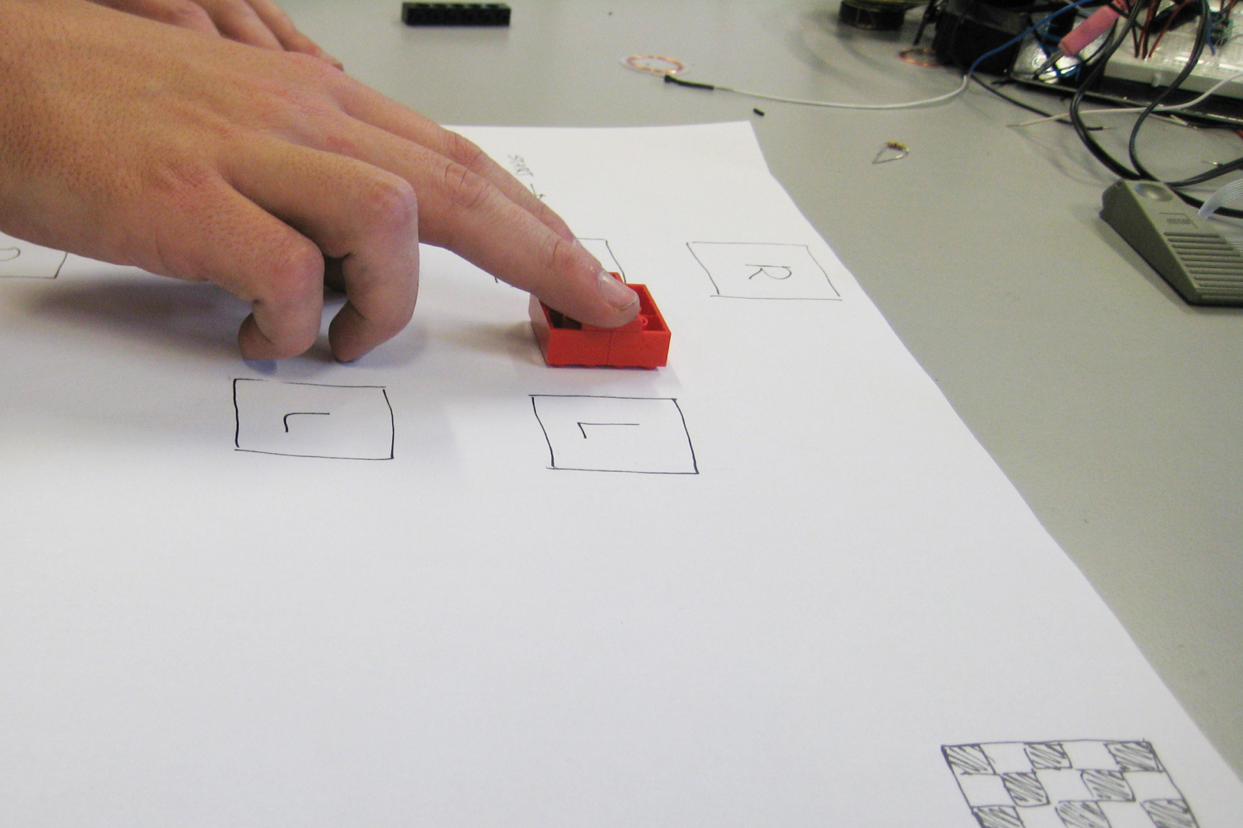 A paper prototype on a table, with a hand moving red Lego bricks across marked fields.