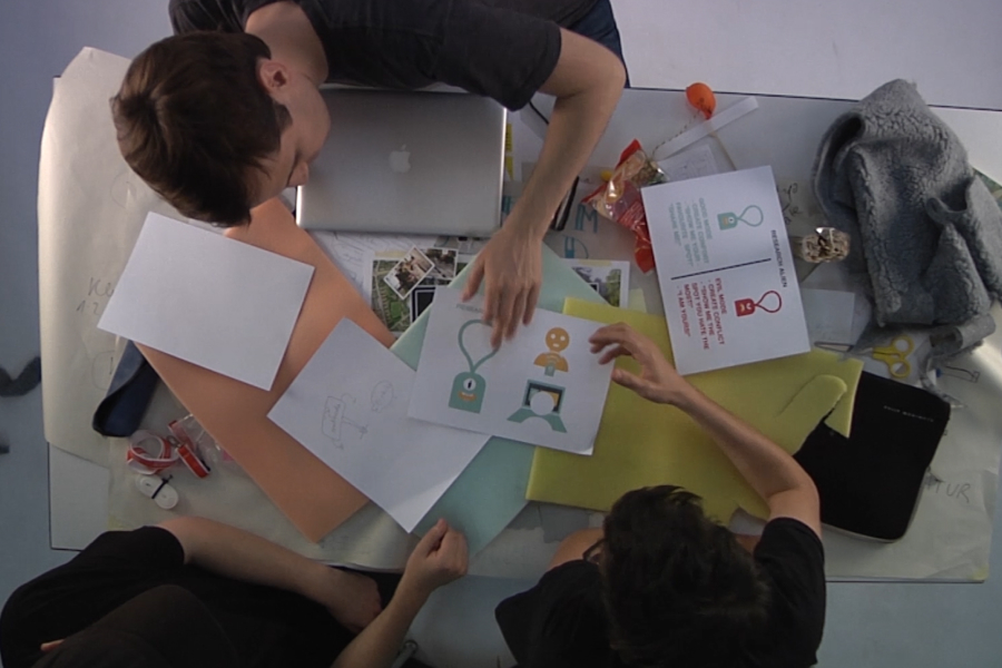Bird's eye view of three students discussing previous findings at a table with design sketches, paper and craft materials.