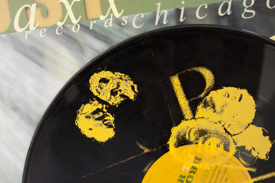 Close-up of a black vinyl record with yellow jazz graphic print, half sticking out of a green and gray patterned record sleeve with typography.
